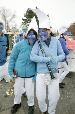 Zwei Personen in blauer Gesichtsbemalung und weißen Kostümen stehen zusammen, eine hält eine Klarinette und die andere eine Trompete. Sie scheinen Teil einer Marschkapelle zu sein.