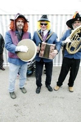 Drei Männer in farbenfrohen Kostümen, jeder mit einem Musikinstrument, stehen in einer Außenumgebung zusammen. Der Mann links hält eine Trommel, der in der Mitte ein Akkordeon und der rechts eine Tuba. Sie scheinen bereit für eine Aufführung zu sein.