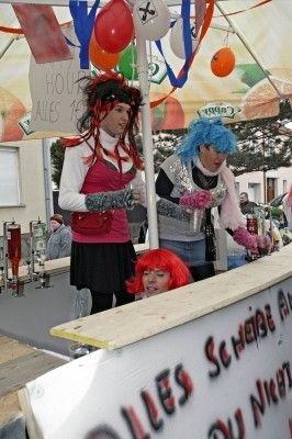 Drei Personen in Kostümen stehen unter einem Zelt mit Luftballons und einem Banner. Eine Person hat rote Haare und schaut in die Kamera. Eine andere Person hat blaue Haare und hält ein Glas. Das Banner steht für 'ALLES SCHONE'.