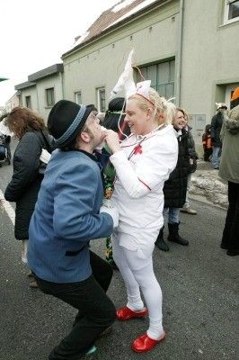 Ein Mann in einem blauen Anzug und Hut küsst eine Frau in einem weißen Uniform und roten Schuhen während einer Straßenveranstaltung.