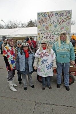 Eine Gruppe von Menschen in bunter und retro Kleidung steht vor einem Friedenszeichen-Banner. Sie posieren für ein Foto.