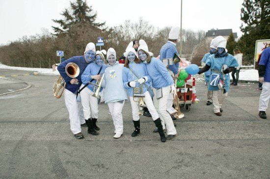 Eine Gruppe von Menschen in blauen Kostümen mit blauer Gesichtsbemalung posiert für ein Foto auf einer Straße. Sie halten Musikinstrumente und Luftballons.
