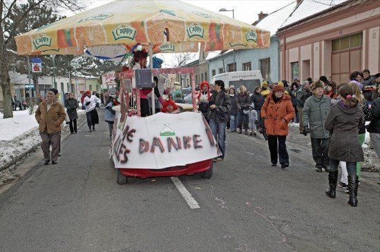 Ein festlicher Umzug mit einem geschmückten Wagen in der Mitte der Straße, um den die Leute herumlaufen. Der Wagen hat ein Baldachin mit dem Wort Cappy und ein Banner mit der Aufschrift 'Die Danke'.