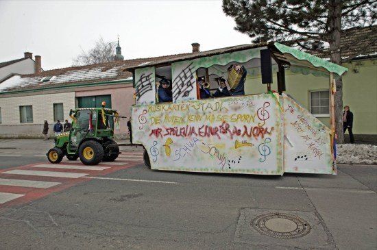 Ein geschmückter Wagen mit Musikern, die Instrumente spielen, steht auf einer Straße, gezogen von einem grünen Traktor. Der Wagen ist mit Noten und Text bemalt.