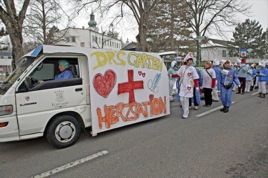 Ein weißer Lieferwagen mit einem Banner, auf dem 'Drs Garten Herzstation' steht, fährt eine Straße entlang, hinter ihm laufen Menschen in medizinischen Uniformen.