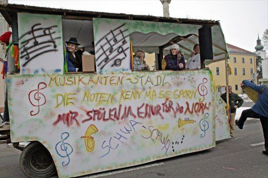 Ein dekorierter Wagen mit Menschen im Inneren, der musikalische Noten und Text in einer fremden Sprache zeigt, der sich in einer Parade mit Gebäuden im Hintergrund bewegt.