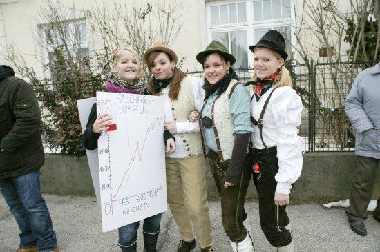 Vier Frauen in traditioneller bayerischer Kleidung stehen zusammen, eine hält ein Schild mit einem Diagramm und Text auf Deutsch. Sie lächeln und scheinen für ein Foto zu posieren. Hinter ihnen ist ein Gebäude mit Glasfenstern und einem Zaun zu sehen.