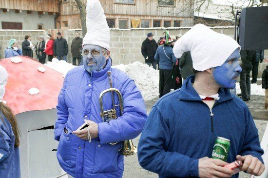Zwei Männer mit blauer Gesichtsbemalung und passenden Mänteln gehen zusammen, einer hält eine Trompete und der andere eine Bierdose. Sie befinden sich in einer verschneiten Außenumgebung mit einer Menge im Hintergrund.