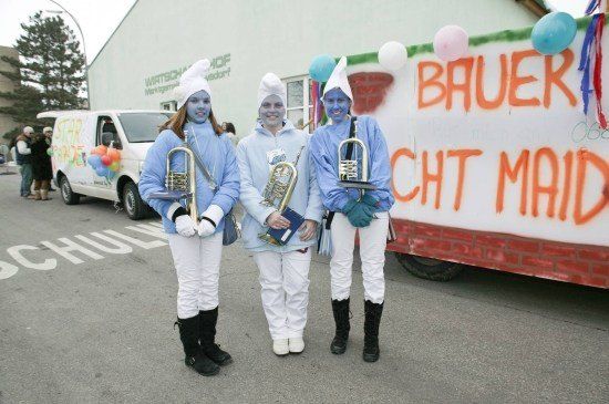 Drei Personen mit blauer Gesichtsbemalung und weißen Kostümen stehen nebeneinander und halten Musikinstrumente in der Hand, mit einem Anhänger hinter ihnen, der 'BAUER ICH MAI' beschriftet ist.