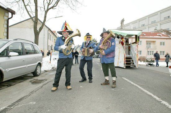 Drei Musiker in bunten Kostümen spielen auf einer verschneiten Straße Instrumente. Ein Auto und ein Gebäude sind im Hintergrund zu sehen.