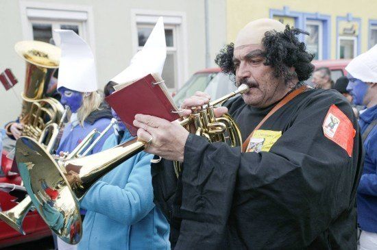 Ein Mann in einem schwarzen Gewand und Turban spielt Trompete und liest Noten, umgeben von anderen mit blauer Gesichtsbemalung und Blechblasinstrumenten.