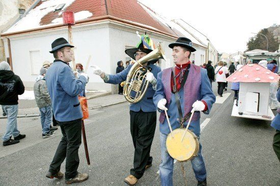 Drei Männer in blauen Kostümen spielen Musikinstrumente auf einer Straße während eines Umzugs. Einer spielt Tuba, ein anderer Trommel, und der dritte hält einen Taktstock. Hinter ihnen ist ein pilzförmiger Wagen und Zuschauer zu sehen.