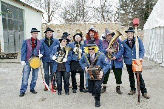 Eine Gruppe von Musikern in farbenfrohen Kostümen, die verschiedene Instrumente wie Trompeten, Posaunen und Trommeln halten, posiert für ein Foto vor einem Gebäude.