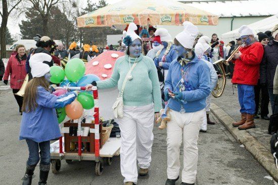 Eine Gruppe von Menschen mit blauer Gesichtsbemalung und weißen Kostümen geht mit einem Korb voller Luftballons. Eine Person in einem roten Mantel spielt ein Blechinstrument.