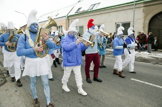 Eine Gruppe von Menschen in blauen Kostümen und weißen Perücken marschiert auf der Straße und spielt Musikinstrumente. Dahinter stehen einige Leute und schauen zu.
