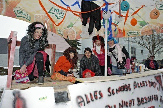 Eine Gruppe von Menschen in Kostümen sitzt auf einer Bühne unter einem Zelt mit einem Banner, das 'Alles Scheine Alles Must' sagt. Sie lächeln und posieren für ein Foto.