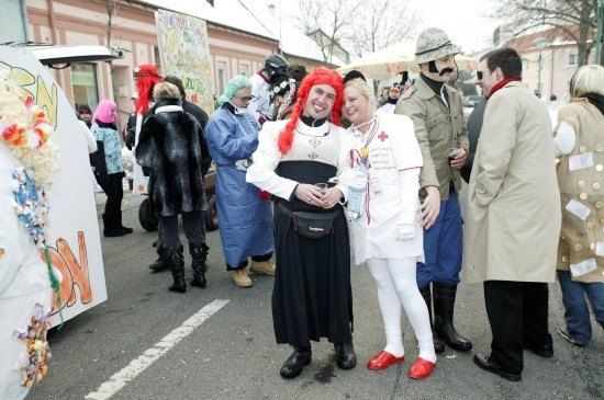 Eine Gruppe von Menschen in verschiedenen Kostümen, darunter eine Krankenschwester und ein Soldat, steht auf einer Straße. Im Hintergrund befinden sich Gebäude und Bäume.