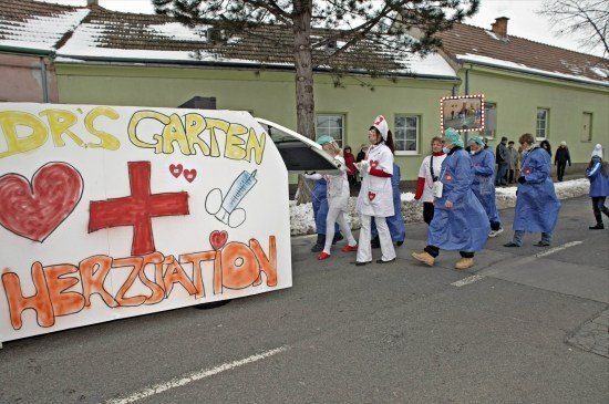 Eine Gruppe von Menschen in blauen Kitteln und weißen Mützen geht bei einer Parade. Ein Banner mit den Worten 'Dr.'s Garten Herzstation' ist am Straßenrand angebracht.