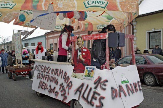 Ein Karnevalswagen mit einem Baldachin und einem Banner mit der Aufschrift 'Alles Danke' steht auf der Straße. Mehrere Leute stehen darum, einige in Kostümen. Ein Auto ist in der Nähe geparkt.