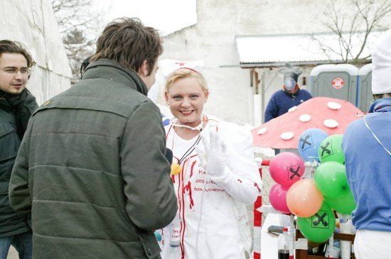Eine Frau in einer Krankenschwester-Verkleidung mit roten Flecken spricht mit einem Mann in einer grünen Jacke. Dahinter steht ein Mann mit Gasmaske bei einem Tisch mit Luftballons.