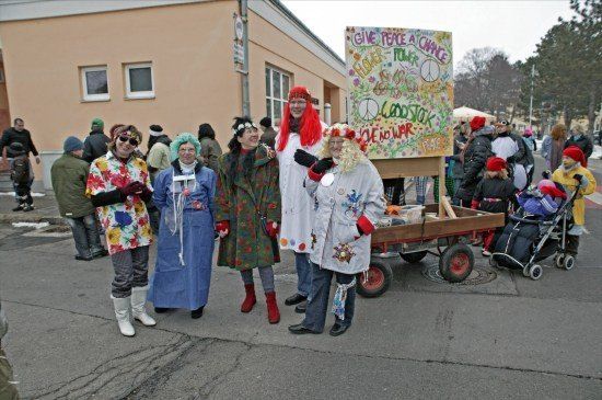 Eine Gruppe von Menschen in bunten Kostümen und Stiefeln steht vor einem Gebäude. Sie halten ein Schild mit den Worten 'Give Peace a Chance' und 'Woodstock'. Ein Kinderwagen steht in der Nähe.