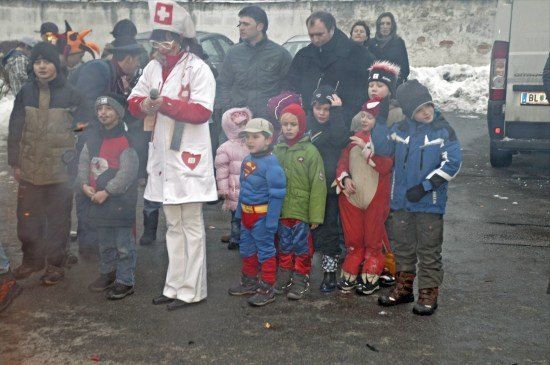 Eine Gruppe von Kindern in verschiedenen Kostümen, darunter ein Superman-Outfit, steht in einer Reihe mit einer Person in weißer medizinischer Kleidung. Die Szene spielt sich an einem verschneiten Außenbereich ab, mit Erwachsenen und einem Van im Hintergrund.