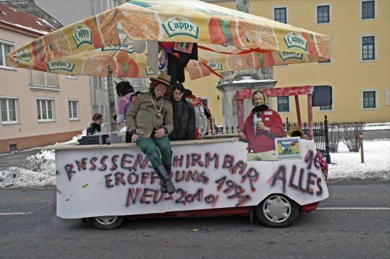 Ein festlicher Wagen mit einem Baldachin, der die Marke Cappy Orangensaft trägt, mit Menschen in Kostümen, einer sitzt auf der Theke, und Graffiti an der Seite mit der Aufschrift 'RIESSEN EROFFNUNG 1993 NEU-2010'.