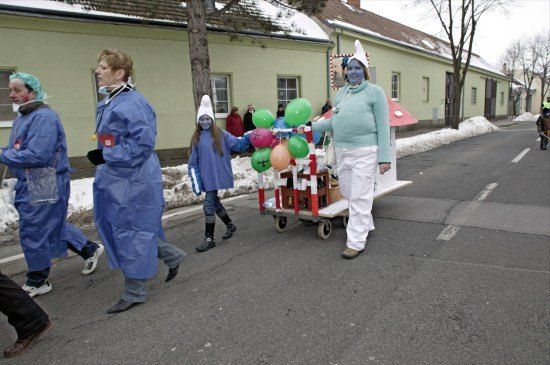 Eine Gruppe von Menschen in blauen Kostümen und Gesichtsbemalung geht eine verschneite Straße entlang, einer schiebt einen Wagen mit Luftballons.