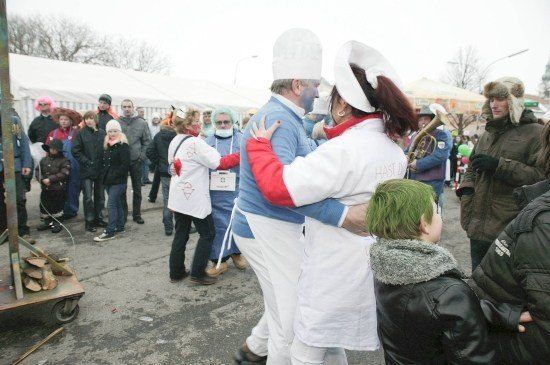 Eine Gruppe von Menschen in weißen Kochuniformen und blauer Gesichtsbemalung tanzt auf einem öffentlichen Platz. Ein Junge mit grünen Haaren beobachtet sie.