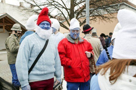 Eine Gruppe von Menschen in verschiedenen Kostümen, einschließlich blauer Gesichtsbemalung und Hüten, steht draußen auf einer Straße. Ein Mann hält eine Trompete.