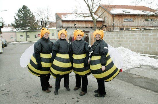 Vier Frauen in Bienenkostümen stehen zusammen in einer verschneiten Außenumgebung. Sie lächeln und posieren für ein Foto. Dahinter befindet sich ein Gebäude mit einer Ziegelmauer und einem Baum.