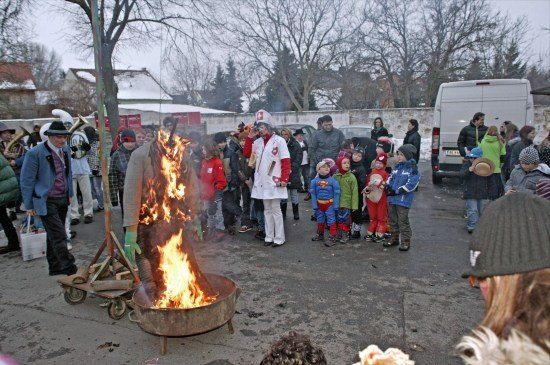 Eine Gruppe von Menschen versammelt sich um ein Lagerfeuer mit einem Mann in einem weißen Mantel und Hut, der davor steht. Einige Leute sind in Kostümen gekleidet und andere tragen Winterkleidung. Ein Van ist im Hintergrund geparkt.