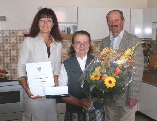 Drei Personen stehen in einer Küche und lächeln. Eine Frau auf der linken Seite hält ein Zertifikat, eine andere Frau in der Mitte hält einen Blumenstrauß und ein Mann auf der rechten Seite trägt einen Anzug.