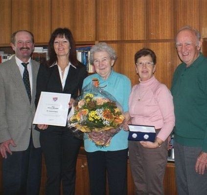 Fünf Personen stehen lächelnd in einem Raum. Eine Frau in der Mitte hält einen Blumenstrauß und ein Zertifikat. Die Frau auf der rechten Seite hält eine gerahmte Auszeichnung. Hinter ihnen sind Holzregale mit Büchern zu sehen.