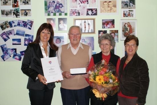 Vier Personen stehen vor einer Wand mit vielen Familienfotos. Eine Frau hält ein Zertifikat und einen Blumenstrauß.