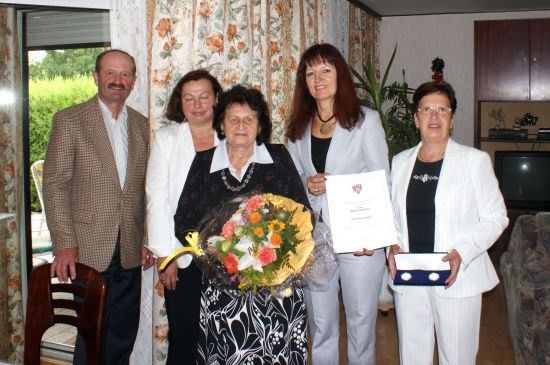 Fünf Personen versammeln sich in einem Raum, eine Frau hält einen Blumenstrauß und ein Zertifikat. Die Frau auf der rechten Seite hält eine Schachtel. Hinter ihnen befindet sich ein Vorhang mit Blumenmustern.