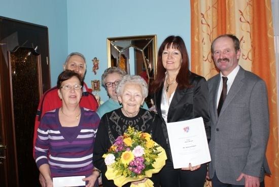 Eine Gruppe von Menschen in einem Raum, eine Frau hält einen Blumenstrauß und ein Zertifikat und posiert lächelnd für ein Foto.