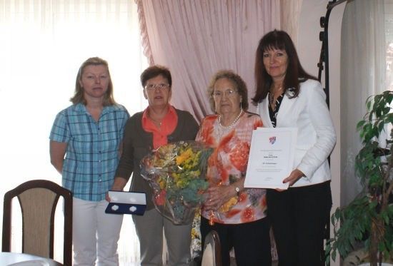 Vier Frauen stehen in einem Raum, zwei halten Urkunden und eine hält einen Blumenstrauß. Die Frau auf der rechten Seite trägt eine weiße Blazer.