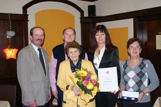 Fünf Menschen stehen lächelnd in einem Raum zusammen. Eine ältere Frau hält einen Blumenstrauß und ein Zertifikat. Eine Frau hält ein Zertifikat und eine Schachtel. Eine weitere Frau hält eine Schachtel. Zwei Männer stehen hinter ihnen.