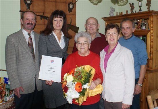 Eine Gruppe von Menschen posiert für ein Foto, wobei eine ältere Frau einen Blumenstrauß und ein Zertifikat in der Hand hält. Hinter ihr stehen ein Mann und eine Frau, und ein weiterer Mann steht rechts. Im Hintergrund befindet sich ein hölzerner Schrank mit verschiedenen Gegenständen.