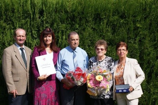 Fünf Personen stehen lächelnd vor einem grünen Busch, halten Blumen und ein Zertifikat.