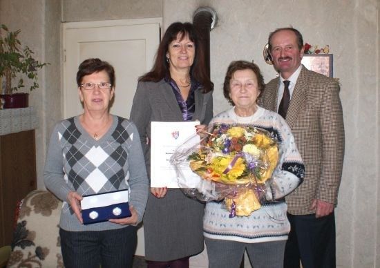 Vier Menschen stehen in einem Raum zusammen, zwei Frauen halten einen Blumenstrauß und ein Zertifikat und ein Mann hält eine kleine Schachtel.
