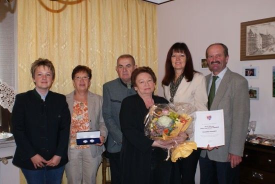 Eine Gruppe von Menschen steht in einem Raum und lächelt für ein Foto. Eine Frau hält einen Blumenstrauß und ein Zertifikat, während ein Mann eine kleine Schachtel hält. Hinter ihnen befindet sich ein gelber Vorhang und ein Bilderrahmen an der Wand.