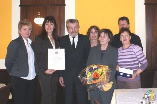 Eine Gruppe von Menschen in formeller Kleidung steht zusammen, lächelt und hält Urkunden und Blumen. Der Mann in der Mitte hält eine Urkunde und einen Blumenstrauß. Hinter ihnen steht ein Tisch mit weißer Tischdecke und einer Lampe.