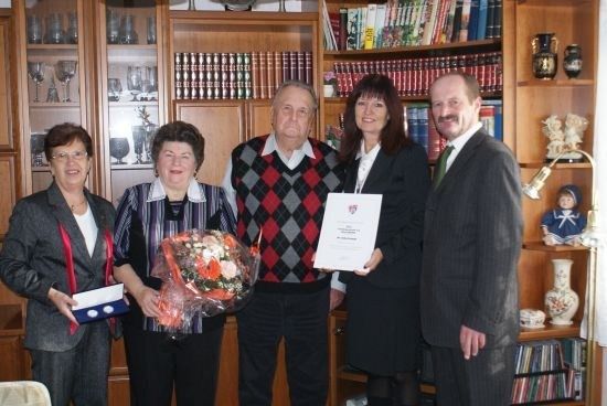 Fünf Personen stehen in einem Raum mit einem Bücherregal im Hintergrund. Der Mann in der Mitte hält ein Zertifikat und einen Blumenstrauß. Die Frau auf der linken Seite hält eine Schachtel. Die Frau auf der rechten Seite hält ein Zertifikat.