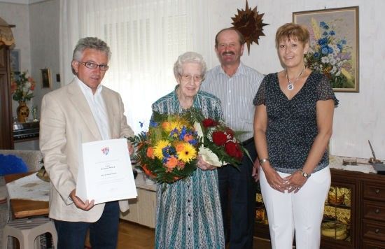 Eine ältere Frau steht zwischen zwei Männern, hält einen Blumenstrauß und ein Zertifikat in der Hand, während eine Frau in einem schwarzen Oberteil neben ihr steht. Sie befinden sich in einem Raum mit einem Holzschrank, einem Tisch mit einer Vase und einem Gemälde an der Wand.