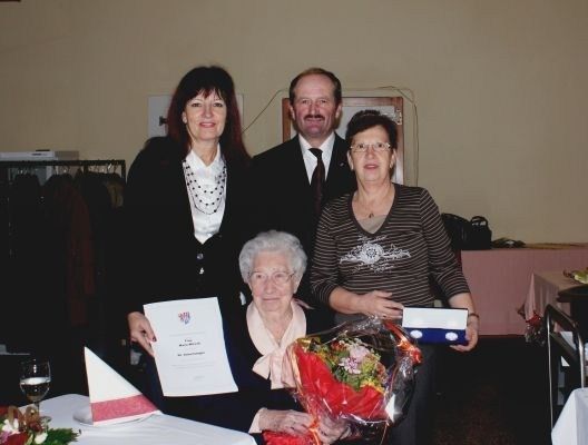 Eine ältere Frau im Rollstuhl wird von zwei Frauen und einem Mann geehrt, die einen Blumenstrauß und ein Zertifikat halten. Sie stehen vor einem Tisch mit einem Weinglas und einer Serviette. Dahinter befindet sich eine Wand mit einem Bilderrahmen und einem Regal mit Kleidung.