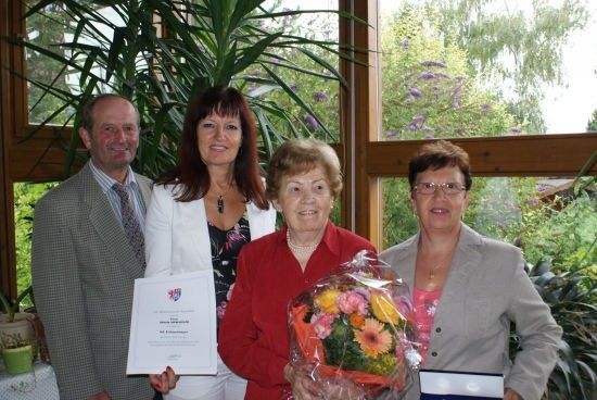 Vier Personen stehen in einem Raum mit Pflanzen und einem Garten im Hintergrund. Die Frau in der Mitte hält einen Blumenstrauß und ein Zertifikat. Der Mann auf der linken Seite hält ein Zertifikat, und die Frau auf der rechten Seite hält ein Zertifikat und eine gerahmte Auszeichnung.