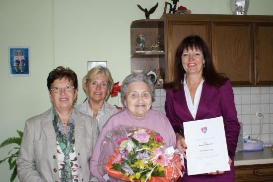 Vier Frauen stehen in einer Küche, eine hält einen Blumenstrauß und eine andere ein Zertifikat. Die Frau mit den Blumen lächelt.