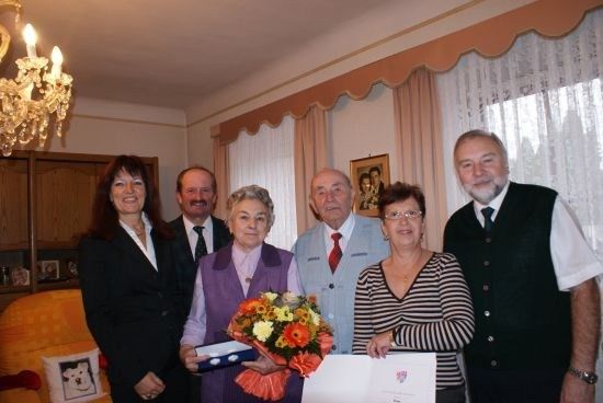 Eine Gruppe von Menschen steht in einem Raum, lächelt und posiert für ein Foto. Eine Frau hält einen Blumenstrauß und ein Zertifikat. Hinter ihnen hängt eine Kristallleuchter von der Decke.